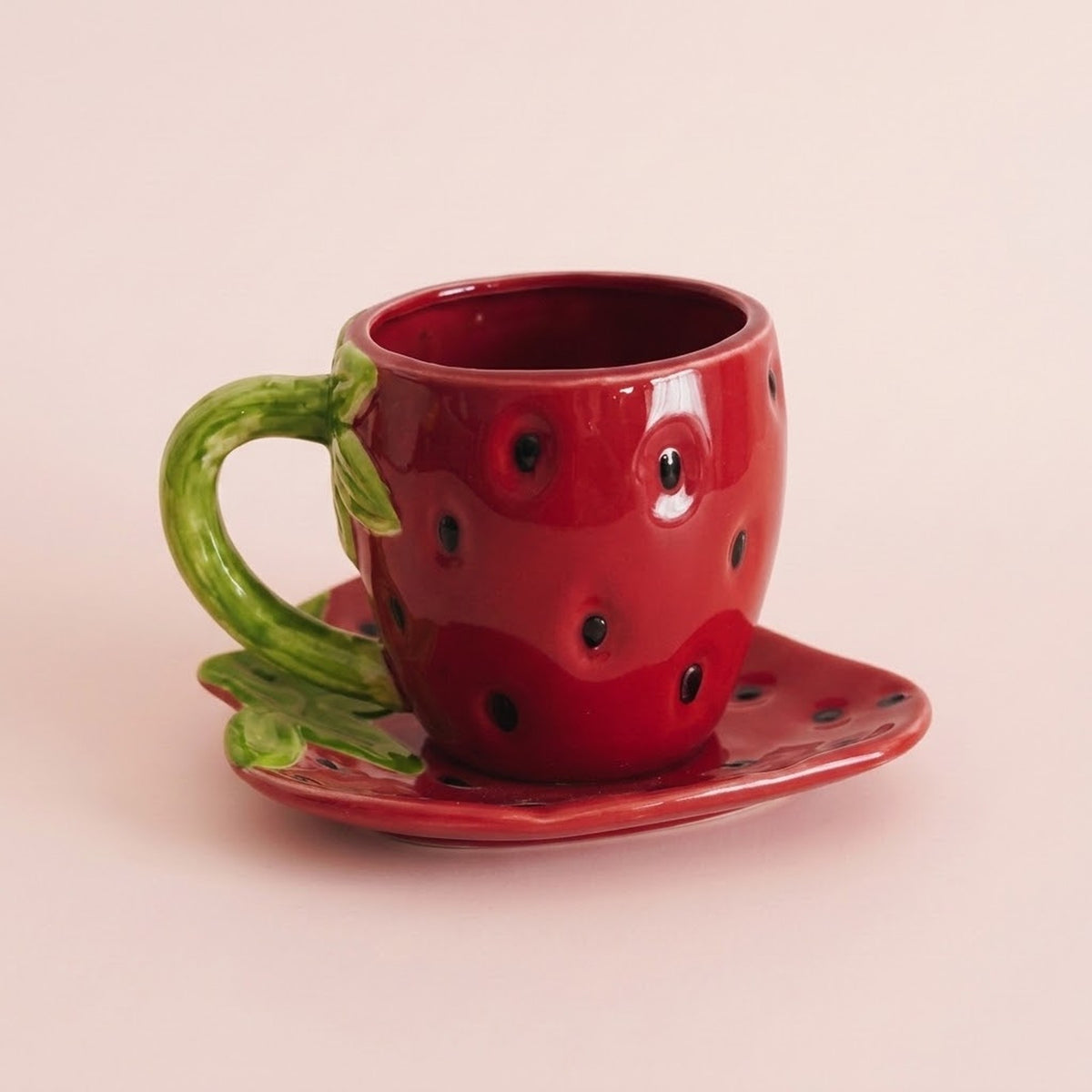 A glossy red ceramic strawberry-shaped cup with a green handle and leaf, shown on a matching saucer against a soft pink background.