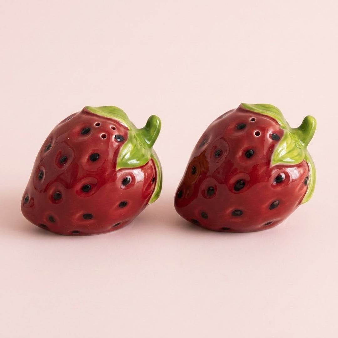Two glossy strawberry-shaped ceramic salt and pepper shakers with green leafy tops, shown on a plain light pink background.