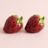 Two glossy strawberry-shaped ceramic salt and pepper shakers with green leafy tops, shown on a plain light pink background.