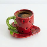 A red ceramic espresso cup designed like a strawberry, filled with black coffee, sitting on a matching red and green strawberry-shaped saucer against a plain white background.