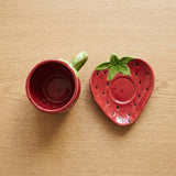 A strawberry-themed ceramic cup and saucer set on a light wood tabletop. The cup is red with a green handle, and the saucer is shaped like a strawberry with a green leafy top.