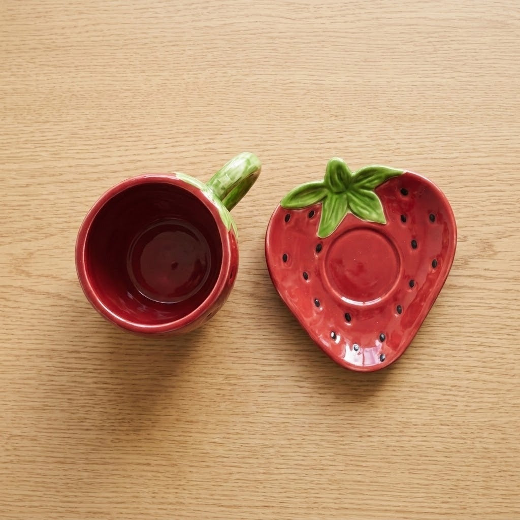 A strawberry-themed ceramic cup and saucer set on a light wood tabletop. The cup is red with a green handle, and the saucer is shaped like a strawberry with a green leafy top.