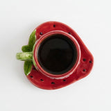 A strawberry-shaped ceramic cup and saucer set, shown from a top-down view. The cup is filled with a dark beverage, and the saucer features small black polka dots and a green handle and stem.