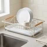 A white wire dish drying rack with light wood handles, shown on a kitchen counter with plates, a mug, and a glass jar, next to a sink and tiled wall.