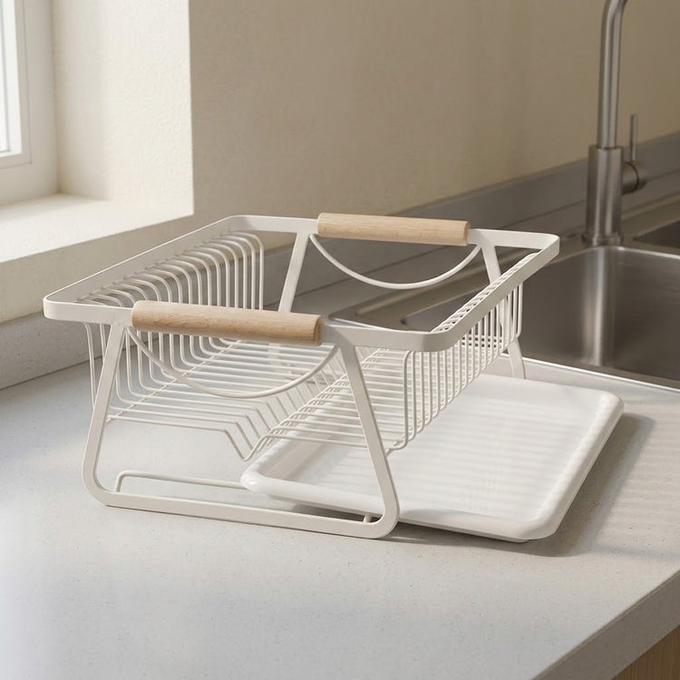 A white wire dish drying rack with light wood handles and a matching white tray, placed on a kitchen countertop near a sink.