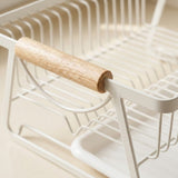 Close-up of a white wire dish rack with a light natural wood handle, set against a warm beige background.
