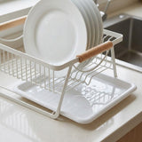 A white wire dish drying rack with light wood handles, shown holding stacked white plates on a kitchen countertop near a sink.