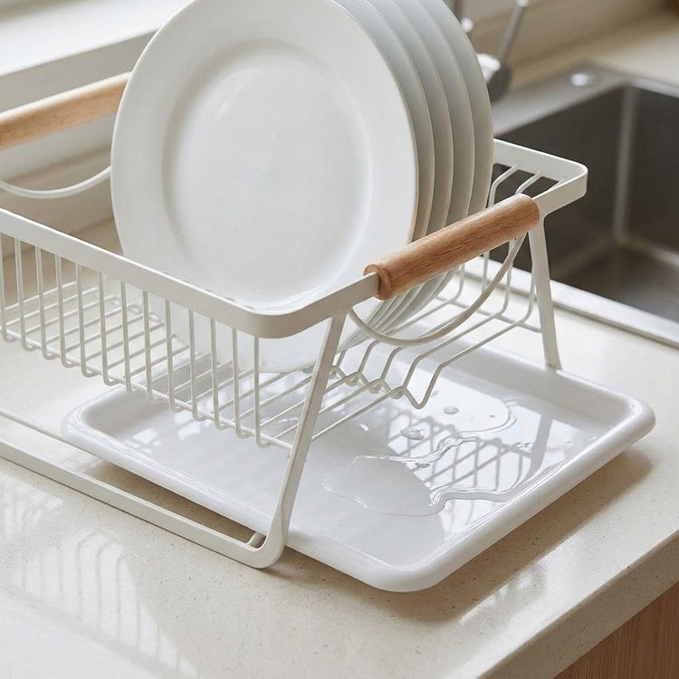 A white wire dish drying rack with light wood handles, shown holding stacked white plates on a kitchen countertop near a sink.