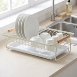 A white wire dish drying rack with light wood handles, shown on a kitchen counter with plates, bowls, and cups inside. A sink and window are visible in the background.