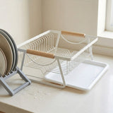 A white wire kitchen dish rack with light wood handles and a white tray, shown on a kitchen counter next to stacked plates.