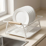 A white wire dish drying rack with a light wood handle, holding several white plates, set on a beige kitchen countertop next to a sink.
