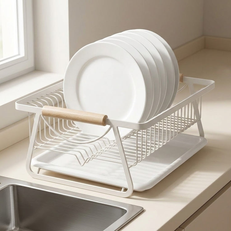 A white wire dish drying rack with a light wood handle, holding several white plates, set on a beige kitchen countertop next to a sink.