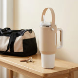 A beige and white insulated travel tumbler with a carry loop and handle sits on a wooden table next to a black and beige duffel bag and a set of keys.