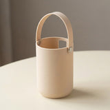 A minimalist, matte beige cylindrical container with a matching arched handle, resting on a light cream-colored table against a soft gray background.