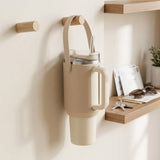 A beige insulated travel tumbler with a carry loop and handle, shown hanging on a wall-mounted holder in a styled interior setting with wooden shelves and small decor items.