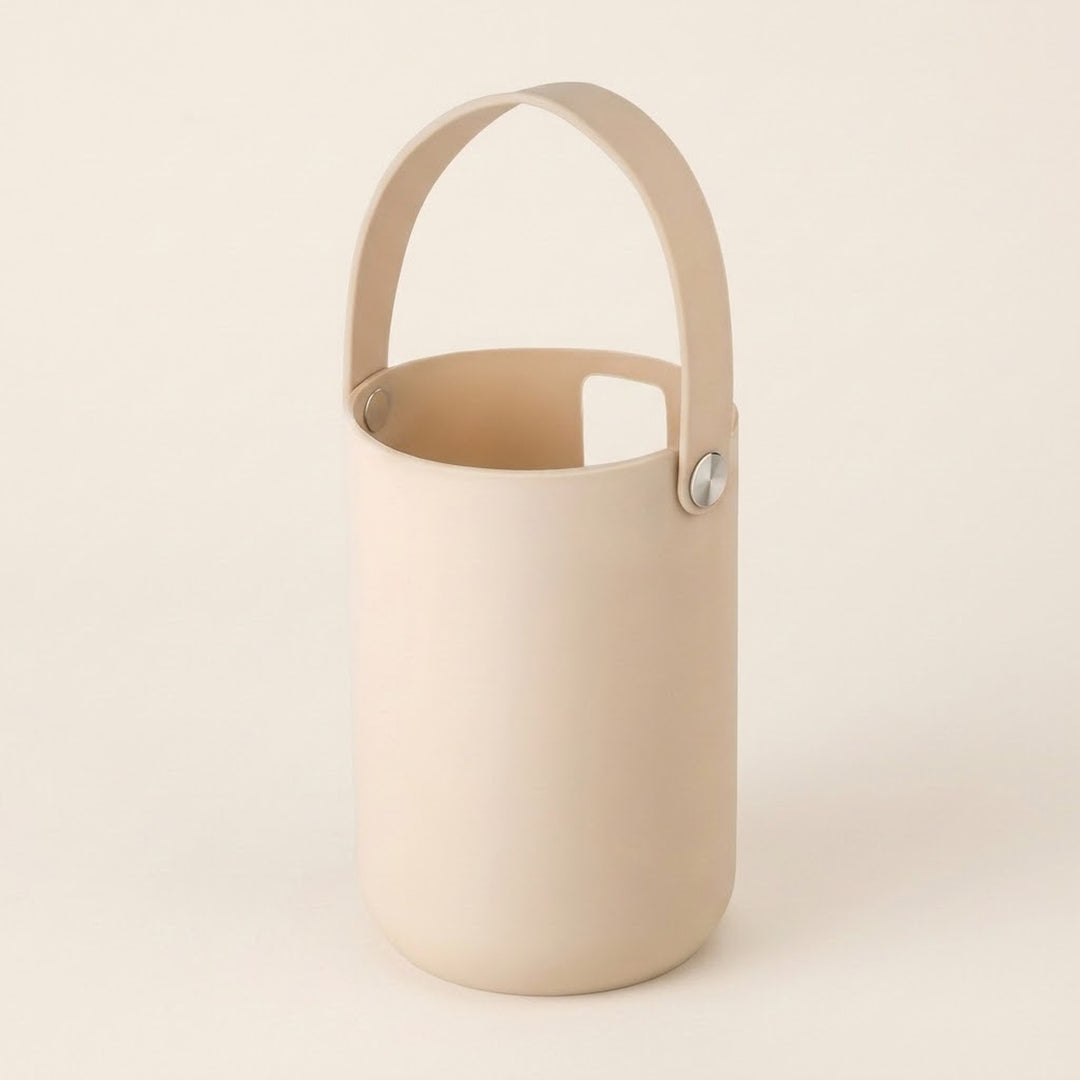 A minimalist cylindrical bucket-style container with a matching top handle, shown on a clean, solid, warm off-white background. The container and handle are a solid cream/beige color, secured with small silver metal rivets.