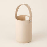 A minimalist cylindrical bucket-style container with a matching top handle, shown on a clean, solid, warm off-white background. The container and handle are a solid cream/beige color, secured with small silver metal rivets.