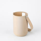 A minimalist light beige leather cylindrical cup with a matching leather handle and a small metal rivet, shown on a seamless white background.