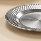 A close-up of a round silver metal plate with a textured, dimpled outer rim and a brushed metallic center, resting on a warm beige surface.