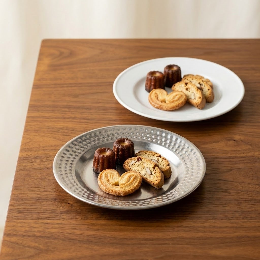 Two ceramic plates with textured metallic rims, one silver and one white, holding assorted cookies and chocolate pieces on a wooden table.