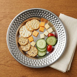 A hammered silver metal serving platter filled with crackers, cheese cubes, cucumber slices, and cherry tomatoes, placed on a wooden table next to a folded cream napkin.