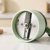 A close-up of a kitchen knife sharpening stone or manual grinder with a pale mint green plastic body and a silver metal sharpening wheel, resting on a light countertop.