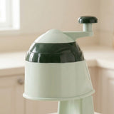A close-up of a countertop kitchen appliance with a pale mint green body and dark green accents, including the lid and handle, set against a softly blurred kitchen background.