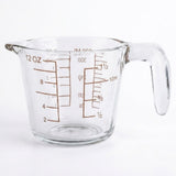 A clear glass measuring cup with a handle, featuring brown printed measurement markings for ounces and milliliters, set against a plain white background.