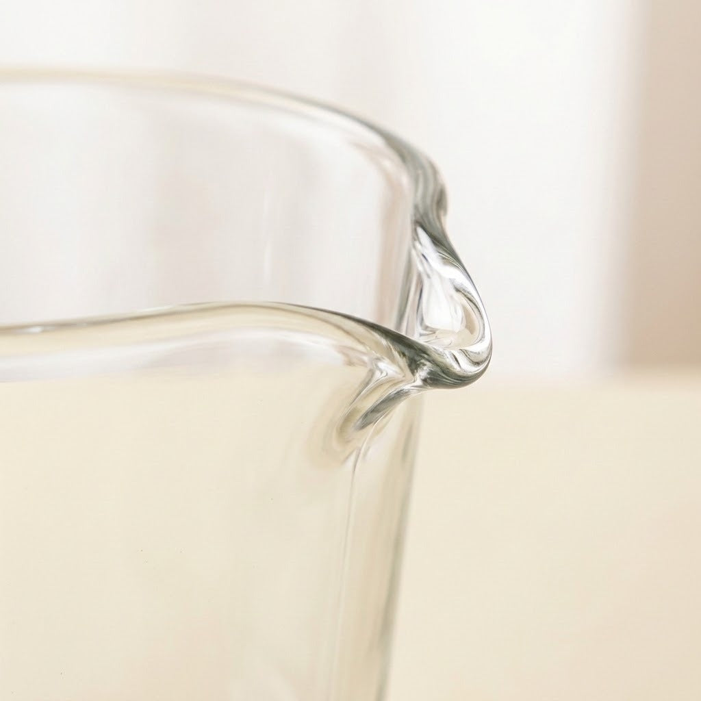 Close-up of a clear glass pitcher with a curved pouring spout, shown against a soft cream-colored background.