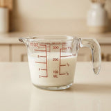 A clear glass measuring cup with red measurement markings, partially filled with white milk, sitting on a light beige countertop in a softly blurred kitchen setting.
