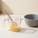 A clear glass measuring cup with red measurement markings holds a yellow liquid and a metal whisk, with a gray ceramic bowl placed behind it on a light beige surface.