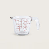 A clear glass measuring cup with red measurement markings and a handle, shown on a plain light beige background.