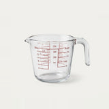 Clear glass measuring cup with red measurement markings and a curved handle, shown on a plain light background.