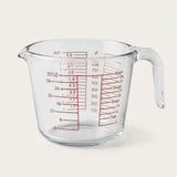 Clear glass measuring cup with red measurement markings and a curved handle, photographed on a plain light background.