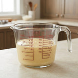 A clear glass measuring cup with red measurement markings, filled with a pale yellow liquid, sitting on a light countertop in a softly blurred kitchen setting.