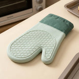 A single oven mitt in pale mint green and dark green rests on a beige countertop, with a blurred toaster oven and baking tray visible in the background.