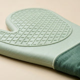 A close-up of a silicone oven mitt in a pale mint green color, showing the textured non-slip grip pattern on the top and a dark green fabric wrist strap, set against a solid beige background.
