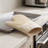 A beige fabric oven mitt with a textured non-slip grip, resting on a stainless steel baking tray on a light beige kitchen countertop next to a built-in oven.