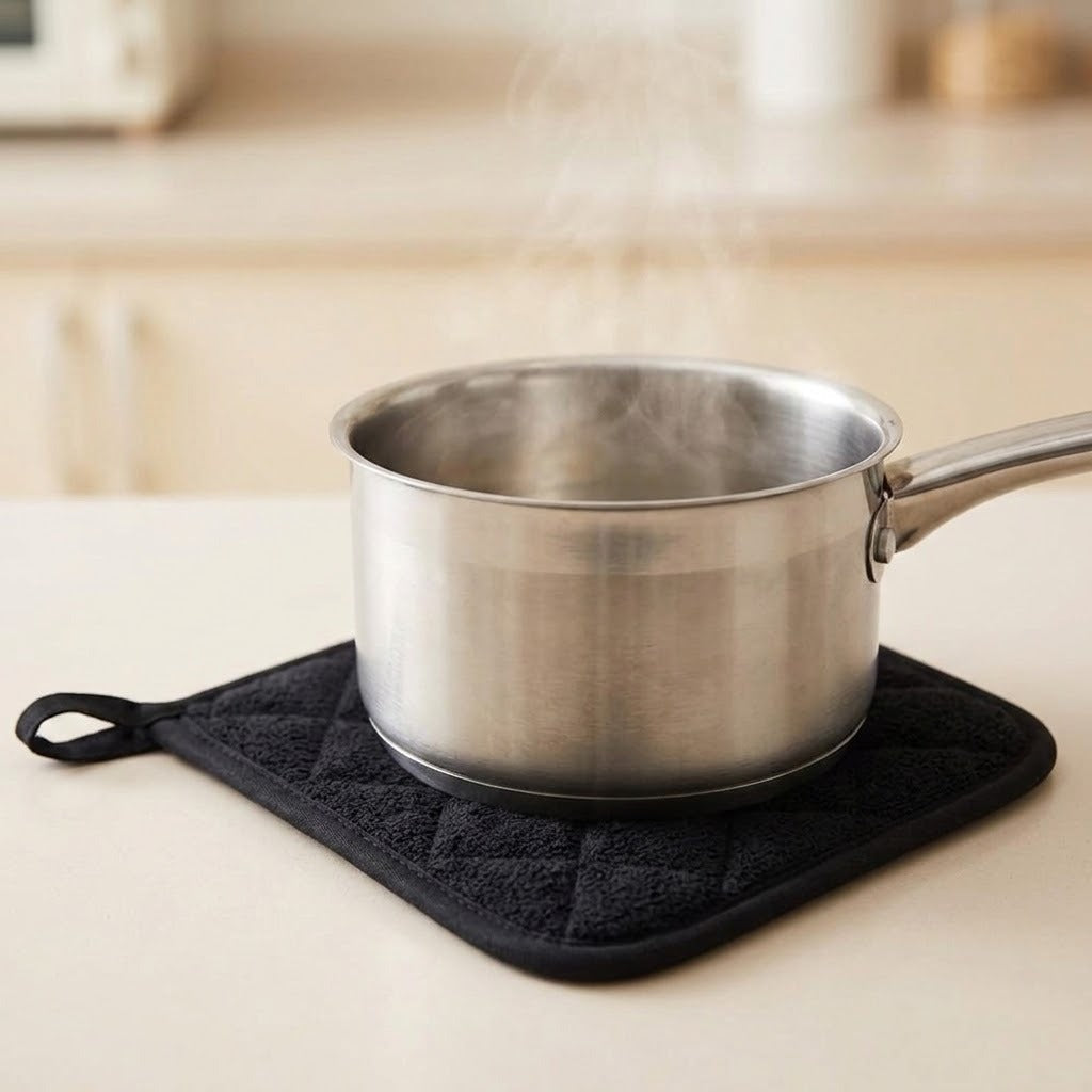 A stainless steel saucepan with a handle sits on a black quilted potholder on a light beige countertop, with steam rising from the pot.