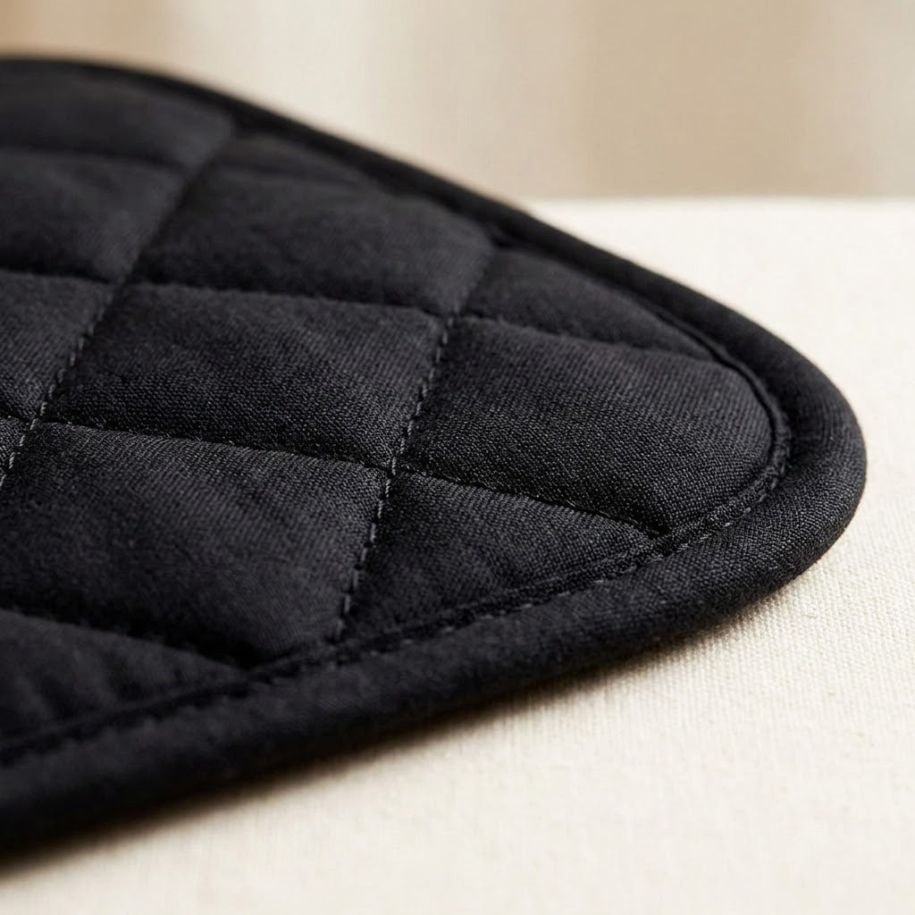 Close-up of a black quilted fabric item, likely an oven mitt or pot holder, showing stitched diamond quilting and a curved reinforced edge on a light neutral surface.