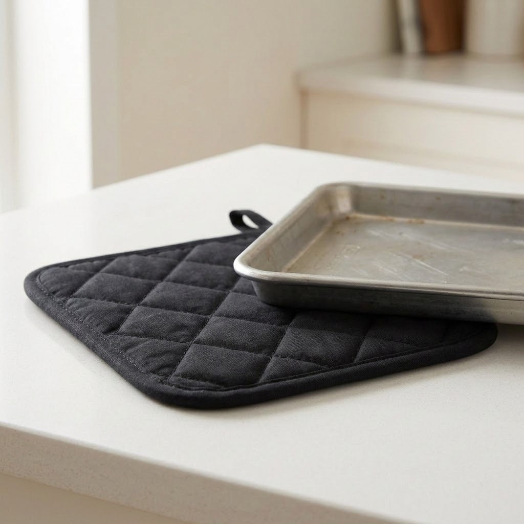 A black quilted oven mitt with a small loop handle rests on a white countertop beneath a metal baking pan, with a softly blurred kitchen background.