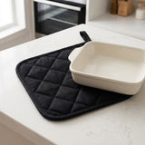 A black quilted oven mitt with a handle resting on a white countertop, with a white ceramic baking dish placed on top.