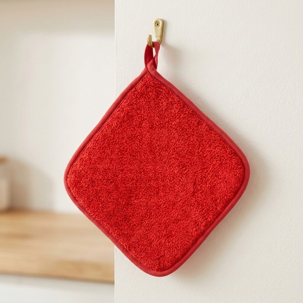 A red textured square potholder with rounded corners and a matching red loop handle, hanging from a small gold hook on a white wall.
