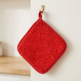 A red textured square potholder with rounded corners and a matching red loop handle, hanging from a small gold hook on a white wall.