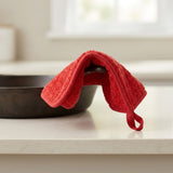A bright red terry cloth kitchen towel with a loop handle draped over a dark cast iron skillet on a light countertop in a kitchen setting.