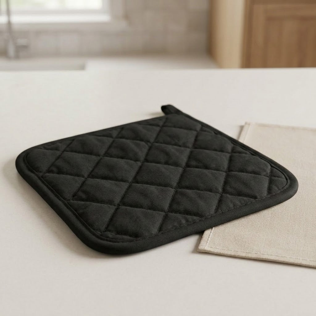 A black quilted oven mitt with a diamond pattern and loop handle, resting on a light-colored countertop next to a beige cloth, with a softly blurred kitchen background.