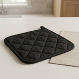 A black quilted oven mitt with a diamond pattern and loop handle, resting on a light-colored countertop next to a beige cloth, with a softly blurred kitchen background.