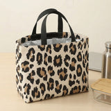 A leopard print insulated tote bag with black handles, shown on a light wood tabletop next to a stainless steel bottle and a glass container with a wooden lid.
