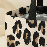 A close-up of a canvas tote bag with a black, beige, and brown leopard print pattern, featuring black webbed handles and a silver foil-lined interior.