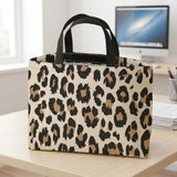 A leopard print tote bag with black handles sits on a light wood desk in front of a blurred computer monitor and office items.